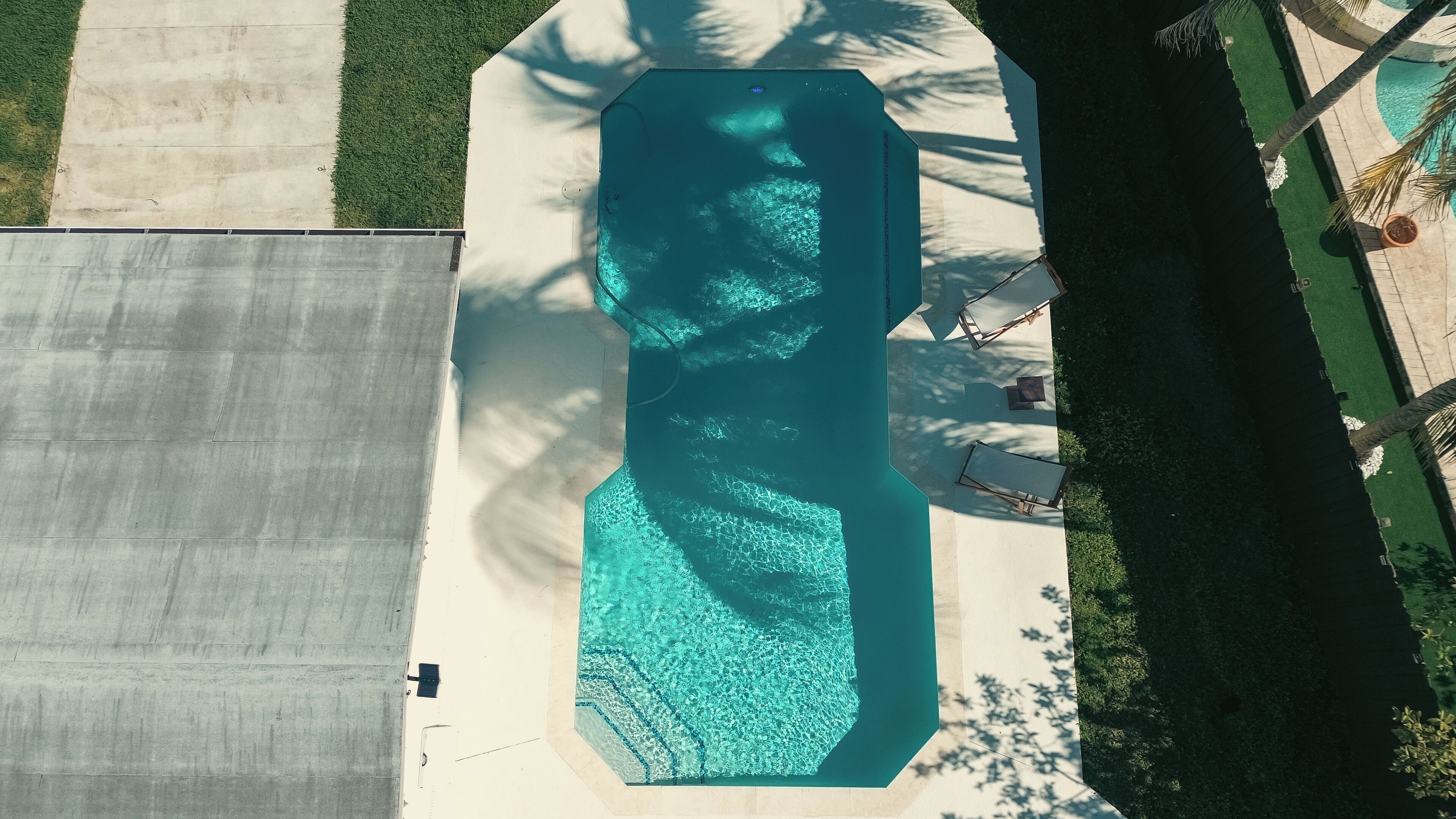 An aerial view of a swimming pool with a geometric shape, surrounded by a concrete deck and palm trees.
