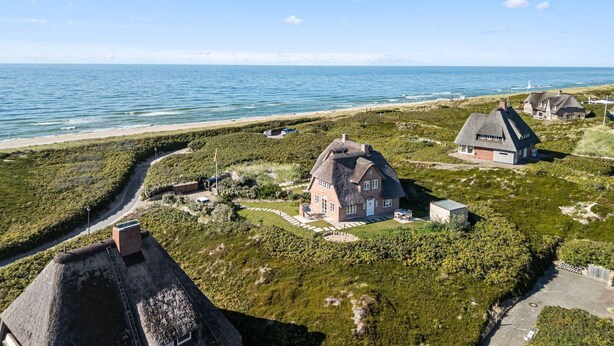 Una propiedad costera con arquitectura tradicional, que incluye una casa principal con techo de paja, una casa más pequeña con techo similar y un faro al fondo.