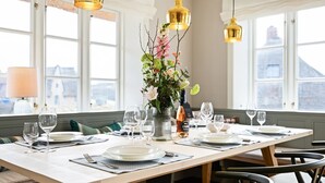 Una mesa de comedor dispuesta con platos, vasos, cubiertos, un florero con flores y una botella de vino, con ventanas amplias y lámparas colgantes al fondo.