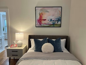 Un dormitorio con cama, una mesita de noche con lámpara, un cuadro abstracto enmarcado y vistas a un balcón.