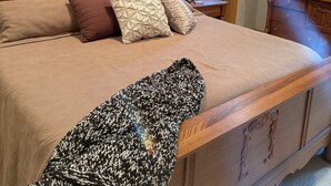 Un dormitorio con una cama de madera, varios almohadones, una colcha, una mesita de noche con lámpara, un aparador y un espejo.