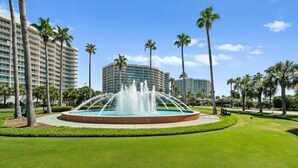 Un hotel con una fuente central, palmeras y un césped bien cuidado.