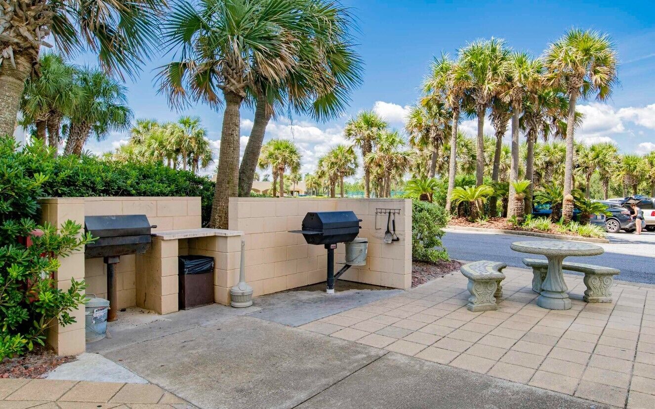 Een terras buiten met barbecuegrills, een stenen tafel en omlijst door palmbomen.