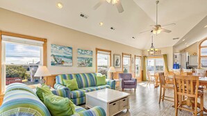 A spacious living area with a large sofa, a dining table, and chairs. There are multiple windows providing natural light and views of the outside.