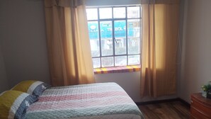 Un dormitori amb llit, terres de fusta, una finestra amb cortines i un ventilador de sostre.