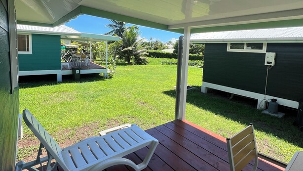 Eine überdachte Terrasse mit zwei weißen Liegestühlen und einem Holzstuhl. Im Hintergrund ist ein Gewächshaus zu sehen.