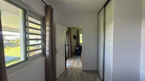 Ein schmaler Flur führt zu einem Schlafzimmer mit einem großen Fenster, einer Deckenleuchte und Blick auf einen Garten.