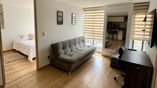 Un salon moderne avec un canapé gris, un plancher en bois, un petit bureau et un coin cuisine à l’arrière-plan.