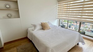 Une chambre avec un grand lit, une fenêtre avec des stores, un plancher en bois et une petite étagère ornée d’objets décoratifs.