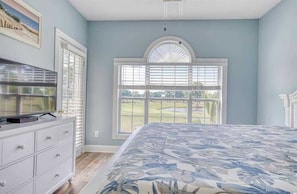 A bedroom with a bed, a dresser, a television, and a large window with blinds.