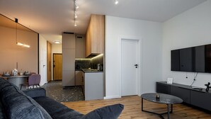 Uma sala de estar moderna com um sofá cinzento, uma mesa de centro e uma área de cozinha com um armário de madeira e uma TV fixada na parede.