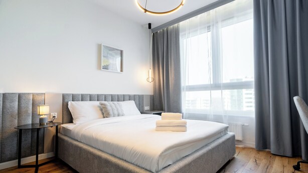 Quarto de hotel moderno com uma cama grande, uma mesinha de cabeceira com abajur, janela com cortinas e vista para a cidade.