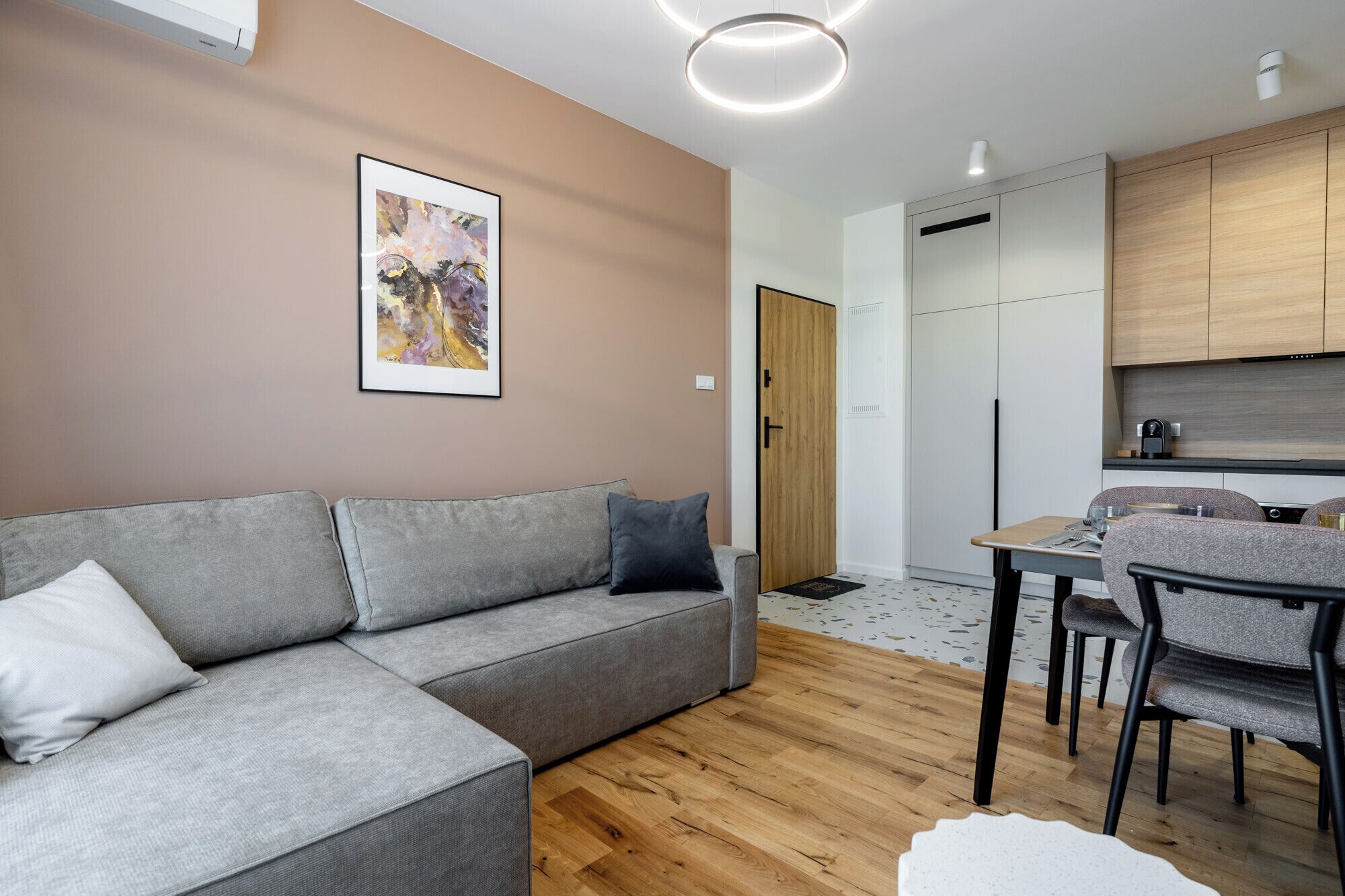 Un soggiorno moderno con un divano grigio, un tavolo da pranzo con sedie e una zona cucina con mobili impiallacciati in legno.