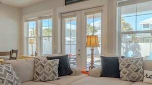 A living room with a beige sofa, patterned cushions, a table lamp, and large windows with blinds.