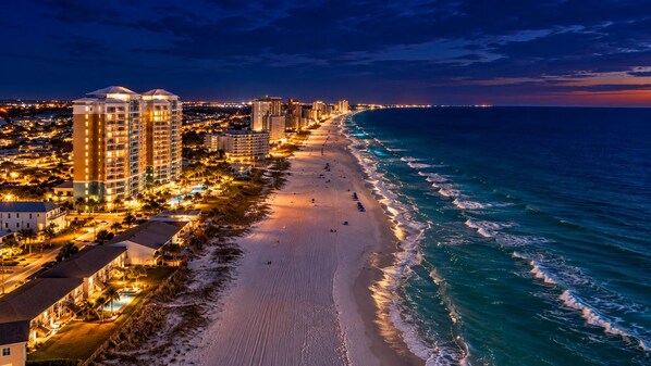 A coastal cityscape at dusk with a beach, high-rise buildings, and a marina.