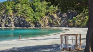 Une plage avec un lit en bois et un auvent, entourée d’une falaise rocheuse et d’arbres.
