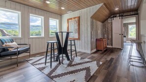 Une pièce spacieuse avec un plancher en bois, une grande fenêtre, une table en métal noir et un tabouret de bar.