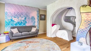 Una sala de estar moderna con un sofá gris, un muro abstracto de colores, una silla de masaje blanca y una alfombra estampada.