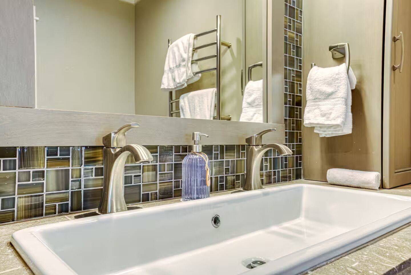 Une salle de bain avec un grand lavabo blanc, deux robinets en chrome, un distributeur de savon en verre et un miroir avec un pare-éclaboussures carrelé.