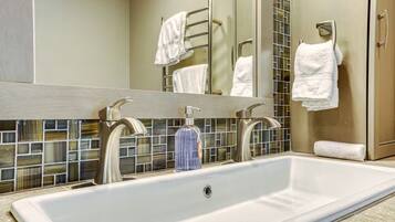 Une salle de bain avec un grand lavabo blanc, deux robinets en chrome, un distributeur de savon en verre et un miroir avec un pare-éclaboussures carrelé.