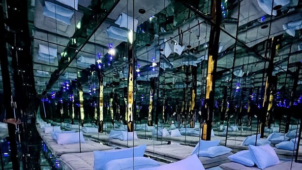 Interior moderno de un hotel con paredes espejadas, varias camas con almohadas y un suelo de cristal.