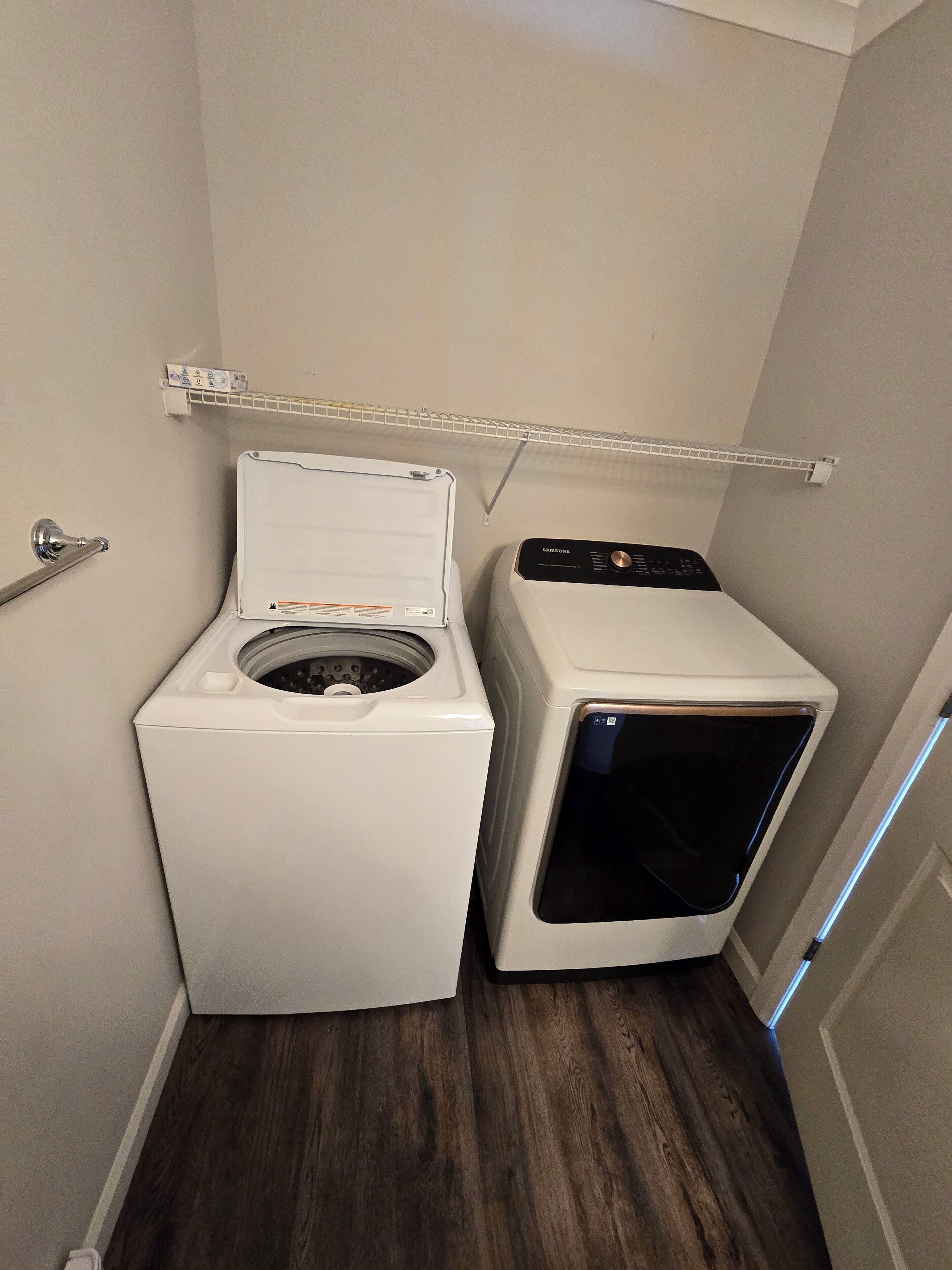 Un lavadero pequeño y funcional con lavadora de carga frontal y secadora de carga superior, ambos blancos, en un espacio reducido con suelo de madera y una estantería metálica arriba.
