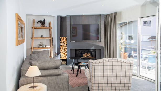 Een moderne woonkamer met een grijze bank, een geruite fauteuil, een bijzettafeltje, een televisie en een open haard met brandhout.