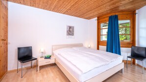 Un dormitorio con techo de madera, una cama con ropa de cama blanca, dos sillas negras, una mesita de noche con una lámpara y una ventana con cortinas azules.