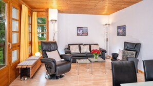 Una sala de estar moderna con techo de madera, un sofá de cuero negro, un sillón de cuero negro, una mesa de centro de vidrio y un banco con cojines.