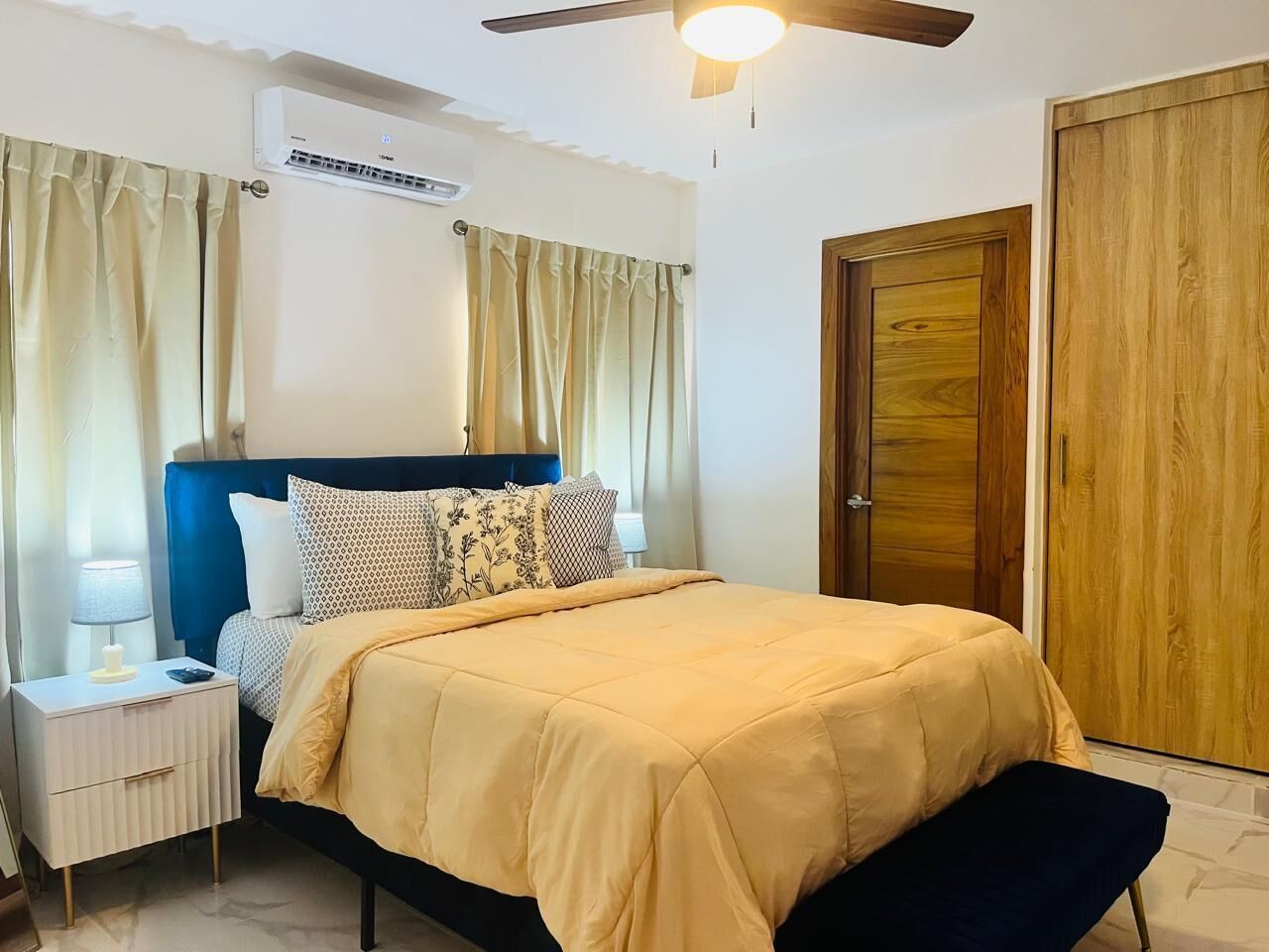 A bedroom with a bed, bedside tables, curtains, an air conditioning unit, a ceiling fan, and wooden doors.