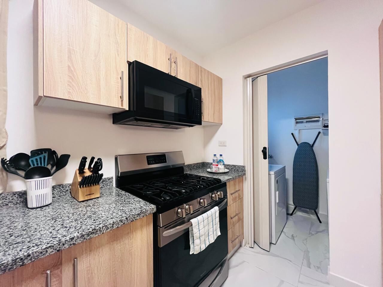 A kitchen with wooden cabinets, a black microwave, a gas stove, and a stainless steel oven. There is a knife block and a bottle of water on the counter.