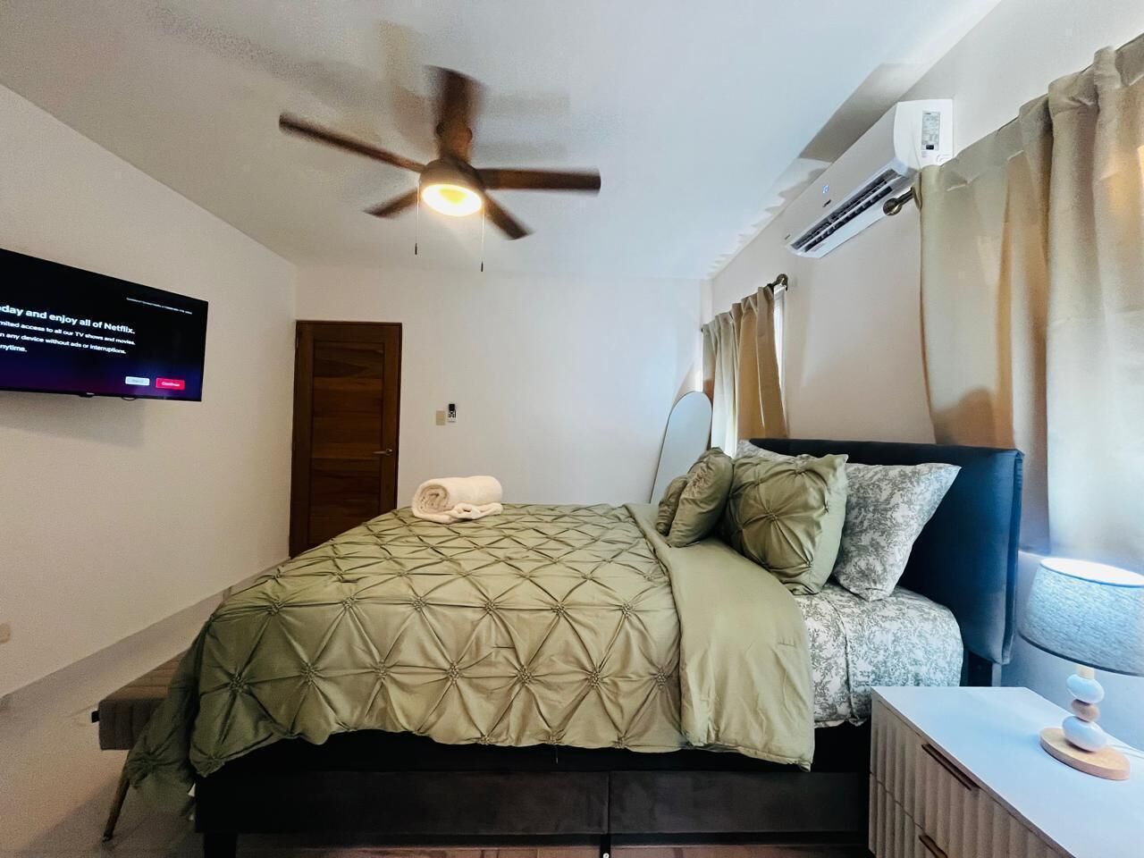 Una camera da letto con un letto, una ventola a soffitto, un condizionatore d'aria, una TV a parete e un comodino con una lampada.