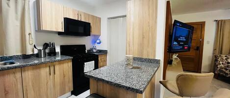 A compact kitchen with wooden cabinets, a microwave, and a granite countertop. There is a small seating area with a chair and a television mounted on the wall.