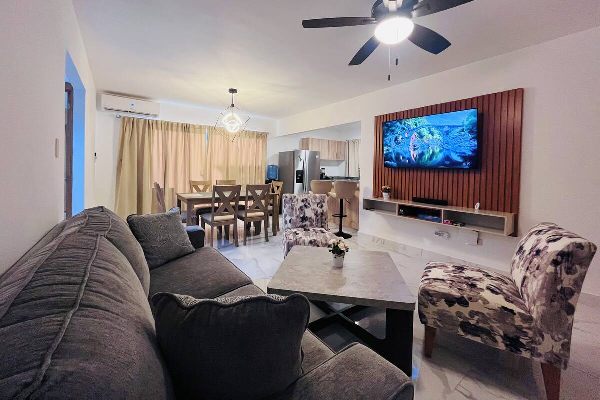 Een moderne woonkamer met een bank, een salontafel en een fauteuil met een patroon. Een plafondventilator en een aan de muur bevestigde televisie zijn zichtbaar.