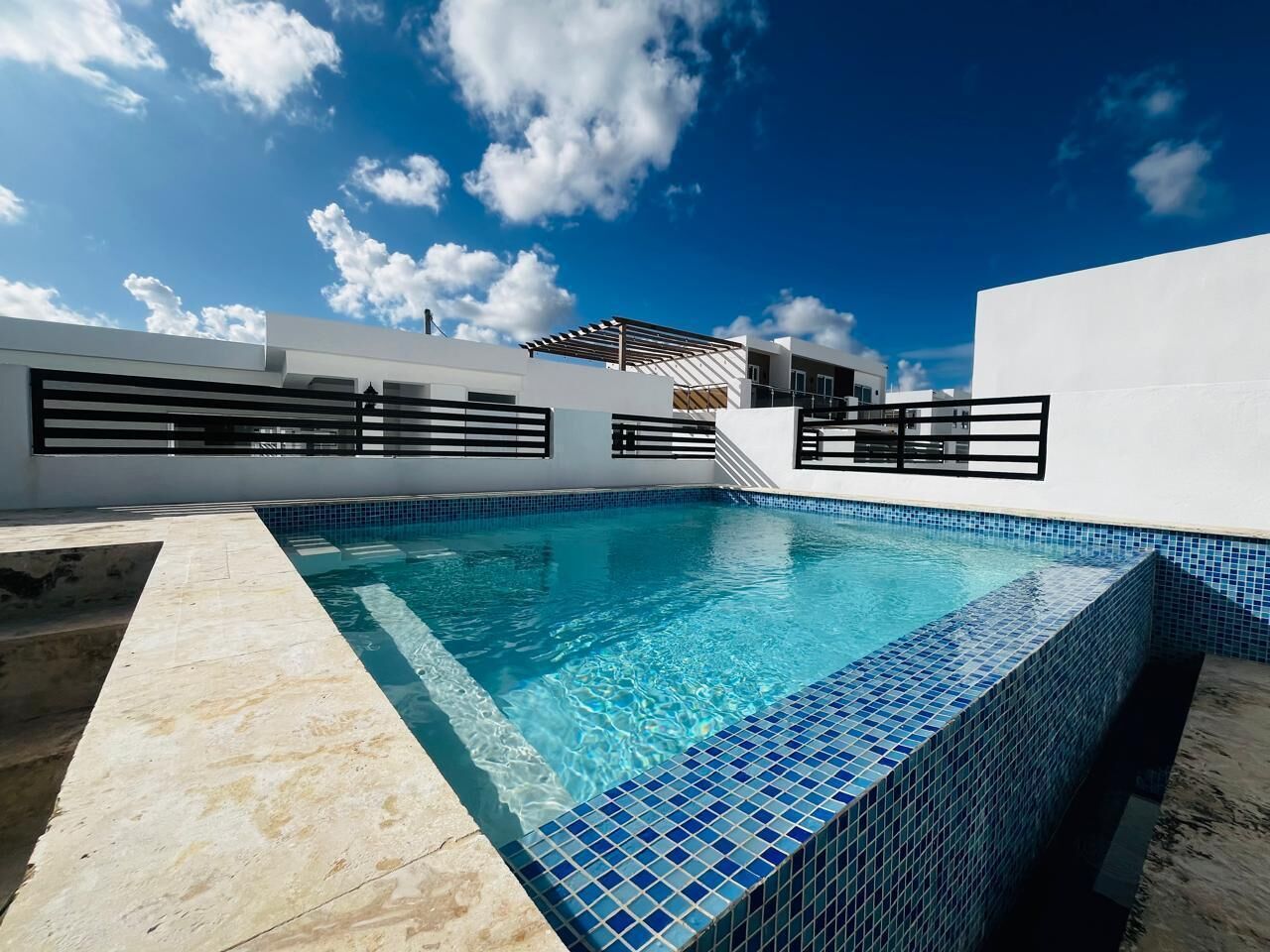 En rektangulær swimmingpool med blå fliser, omgivet af en hvid mur og en pergola.