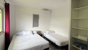 A room with two beds, a window with red curtains, a wall-mounted air conditioner, and a wooden floor.