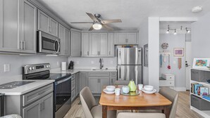 A modern kitchen with grey cabinets, stainless steel appliances, a wooden dining table, and a ceiling fan. - Private Balcony, Ocean View, Steps to the Beach - Sunsets & Sanddollars - Roelens (Fort Myers Beach)