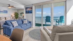 A modern living room with a blue sofa, beige armchair, and a round rug. A large window offers a view of the ocean and a balcony with blue chairs. - Private Balcony, Ocean View, Steps to the Beach - Sunsets & Sanddollars - Roelens (Fort Myers Beach)