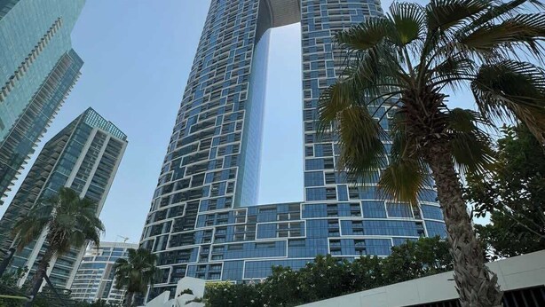Un immeuble moderne de grande hauteur, doté d’une fenêtre verticale originale, et entouré de palmiers et de verdure.