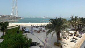 Une vue sur la plage avec une grande roue, des palmiers, et une plage parsemée de parasols et de chaises longues.