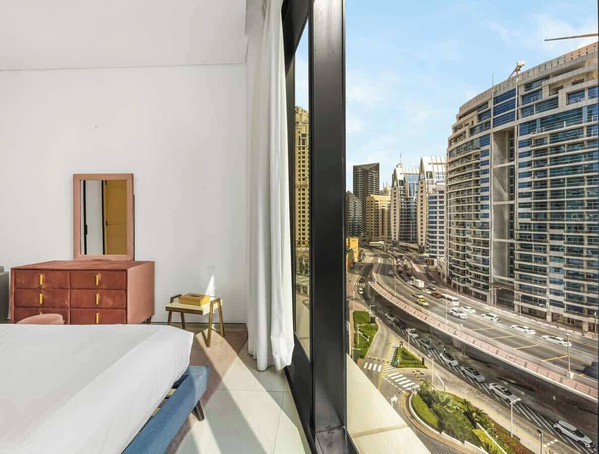 Una habitación de hotel moderna con una cama grande, un aparador, una silla y vistas a una ciudad bulliciosa con edificios altos y una autopista.