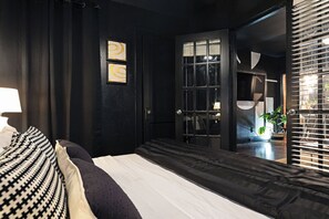 Een slaapkamer met een bed, een raam met zonwering, een deur en een muur met een spiegel en kunstwerk.