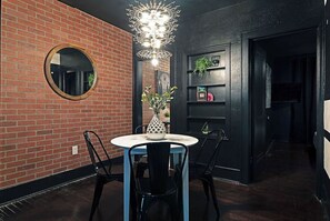 Een eetgedeelte met een bakstenen muur, een ronde spiegel, een kroonluchter, een ronde tafel met stoelen, en een plank met planten.