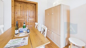 Ein Holztisch mit einer Tischmatte, einer Weinflasche und einer Speisekarte. Weiße Stühle stehen um den Tisch, und im Hintergrund ist ein Holzschrank zu sehen.