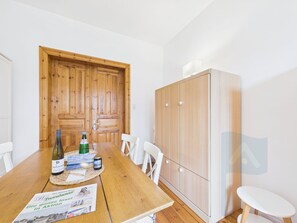 Ein Holztisch mit einer Tischmatte, einer Weinflasche und einer Speisekarte. Weiße Stühle stehen um den Tisch, und im Hintergrund ist ein Holzschrank zu sehen.
