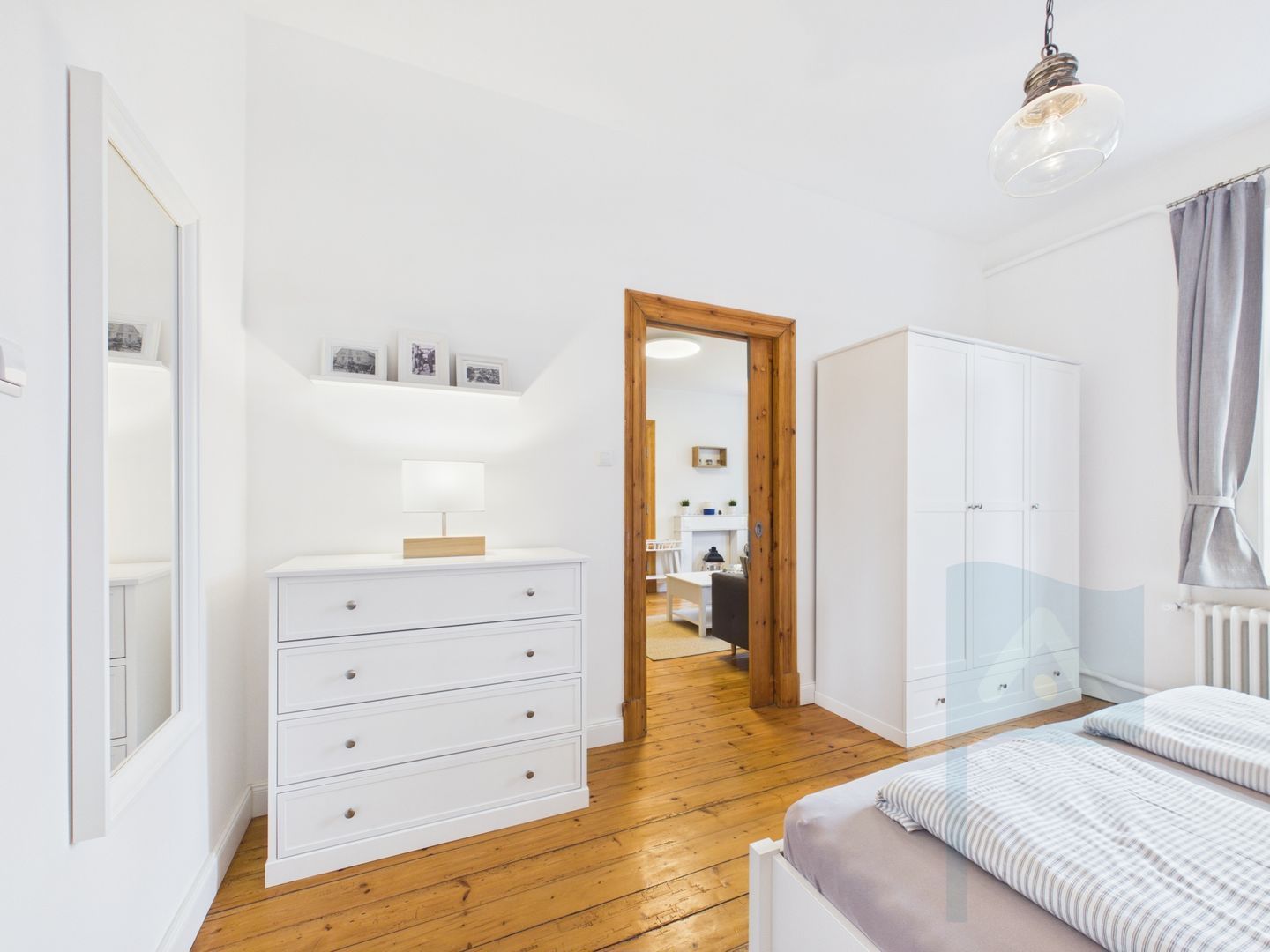 Quarto com piso de madeira, mobiliário branco, cama com roupa de cama listrada, guarda-roupa e mesa de cabeceira com abajur.