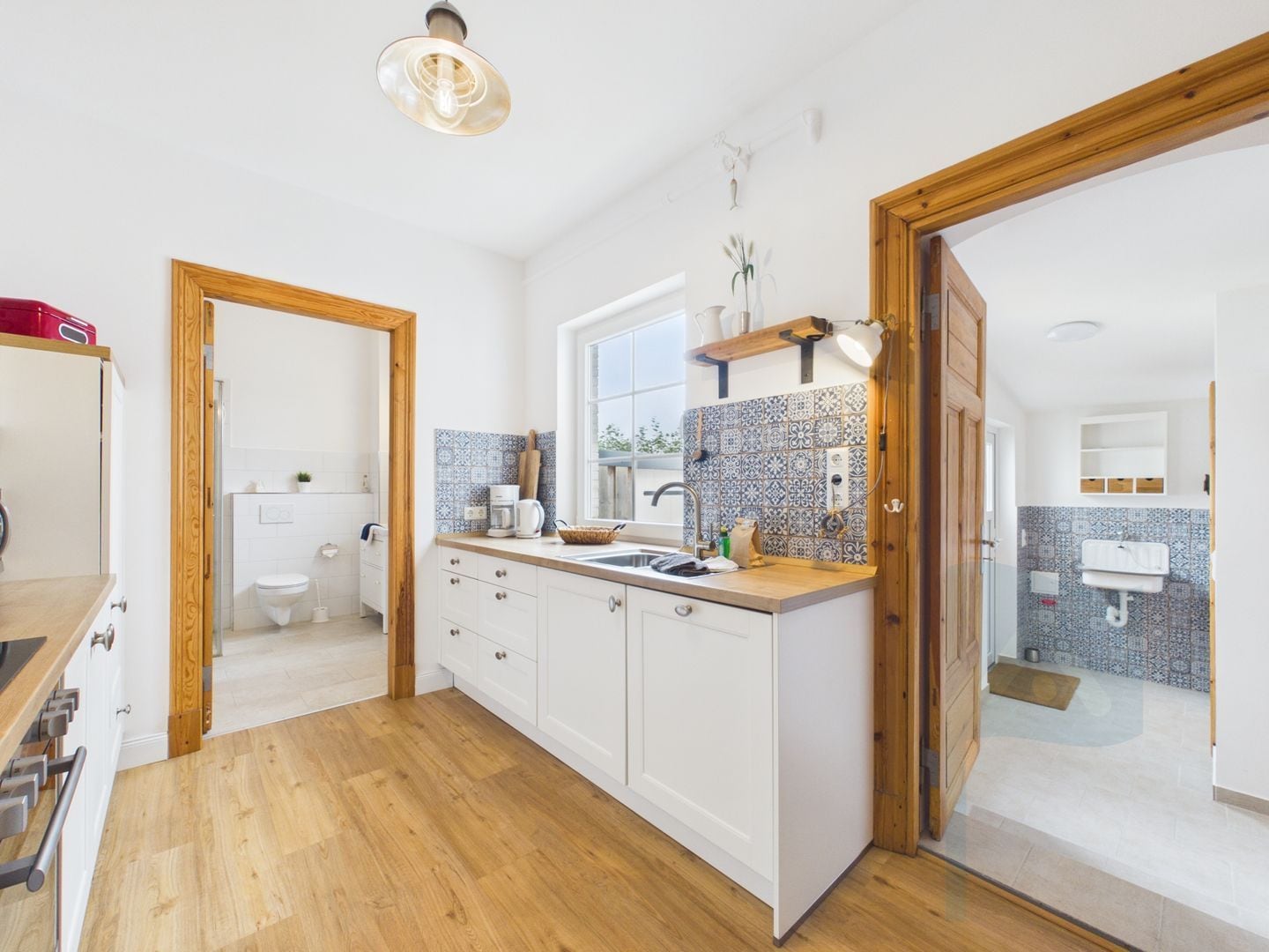 A modern kitchen with wooden flooring, white cabinetry, a large window, and a tiled backsplash.