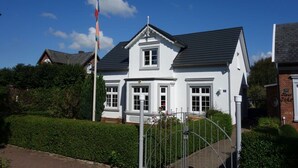 Ein weißes, zweistöckiges Haus mit dunklem Dach, umgeben von Grünpflanzen und einem Metallzaun.