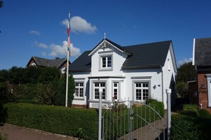 Ein weißes, zweistöckiges Haus mit dunklem Dach, umgeben von Grünpflanzen und einem Metallzaun.
