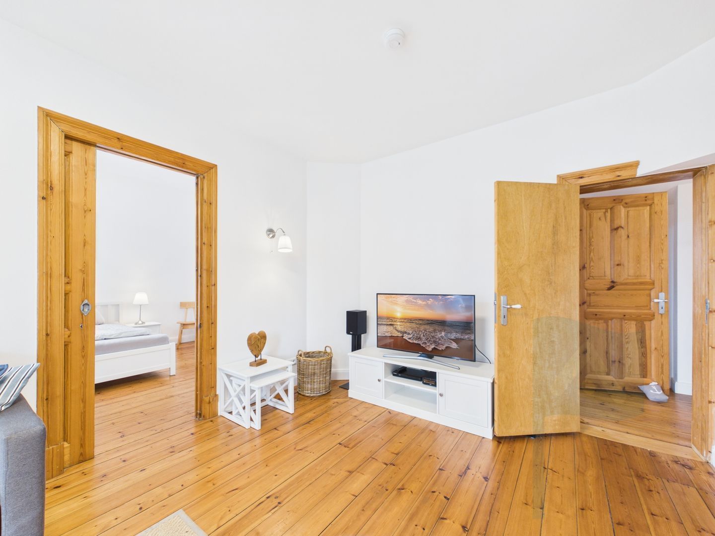 A room with wooden flooring, a television on a white stand, a small white table with a basket, and a door leading to another room.